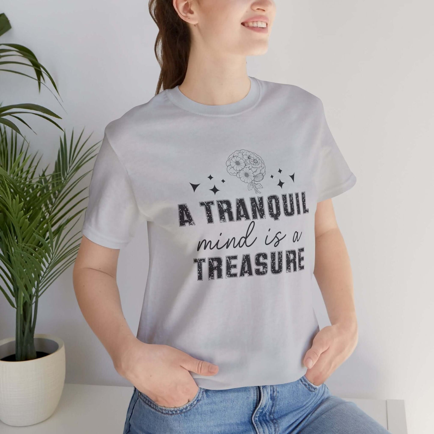 Woman wearing a white t-shirt with 'A tranquil mind is a treasure' floral print, standing indoors.
